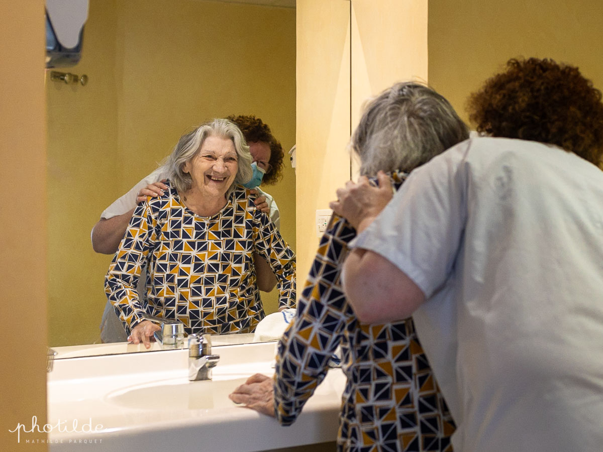 résidente avec une aide-soignante se regardent dans le miroir d'une salle de bain d'un ehpad et sourient 