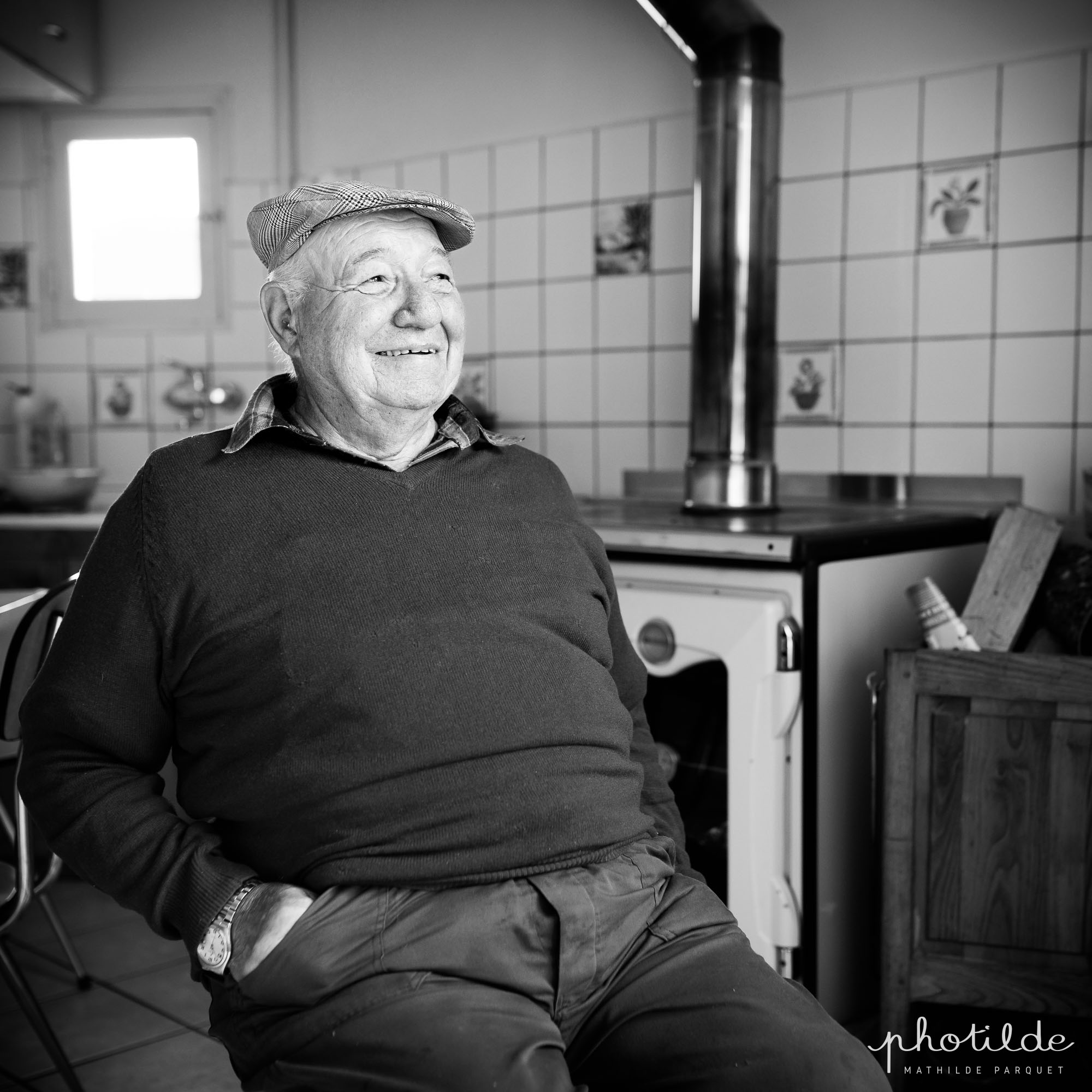 Monsieur de quatre vingt ans environ assis dans sa cuisine. Il porte un casquette et est souriant. une vieille gazinière à bois est en arrière plan.