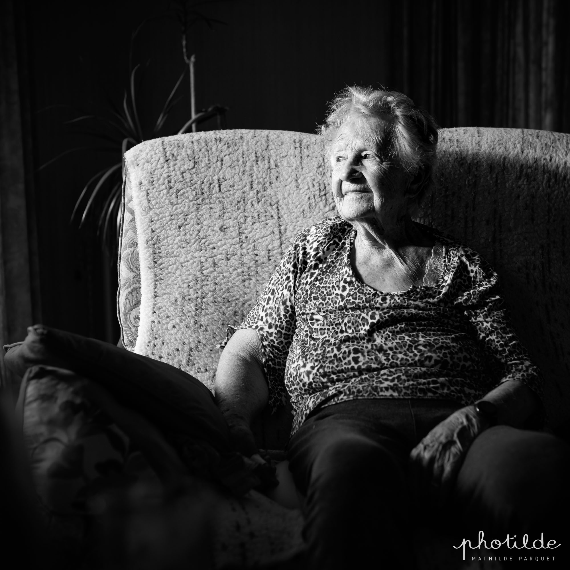 Femme centenaire assisse dans son canapé. Un joli rai de lumière vient l'éclairer, elle regarde vers la lumière.